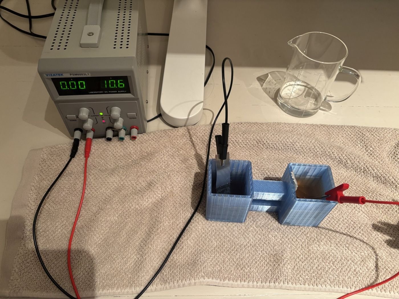 Blue 3D-printed two-chamber cell on a towel, nickel electrodes wired to a bench DC power supply
